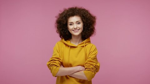 Cheerful happy girl afro hairstyle in hoodie turning crossed hands and looking at camera with toothy smile, expressing confidence optimism, positive emotions. studio shot isolated, pink background