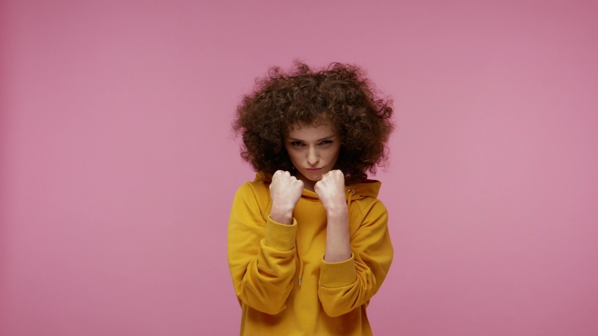 Let's fight! Brave girl afro hairstyle in hoodie punching fists to camera, boxing with frowning aggressive face expression, self defense concept. indoor studio shot isolated on pink background | Shutterstock HD Video #1056818900