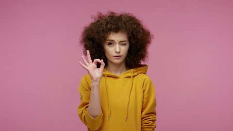 I'm ok! Pretty positive girl afro hairstyle in hoodie showing okay hand sign and winking as if approving suggestion, completely agree and support. indoor studio shot isolated on pink background