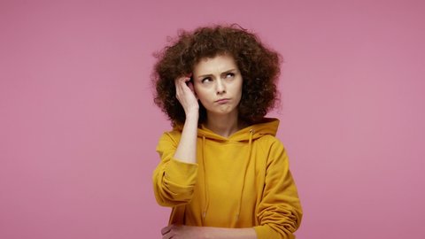 Thoughtful confused girl afro hairstyle in hoodie touching chin, scratching head while thinking over solution, having doubts about difficult choice. indoor studio shot isolated on pink background