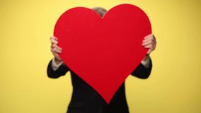happy elegant businessman in suit hiding behind big red heart, showing and presenting heart on yellow background