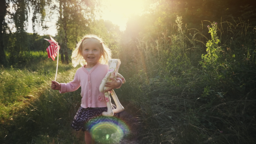 Little smiling girl runs along a path in nature in the rays of sunlight holding a toy windmill and a toy hare in her hands, slow motion