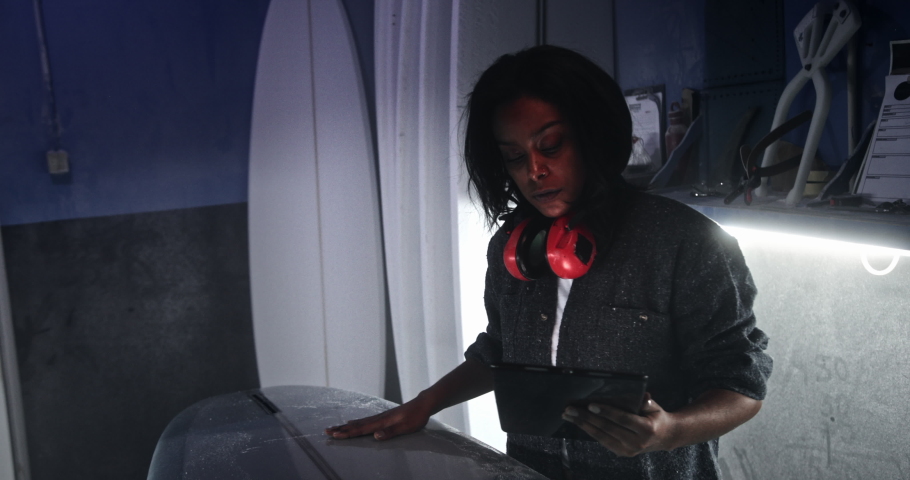 Black woman checking surfboard and working on tablet in workshop