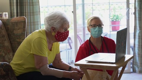 Closeup of an elderly woman and nurse, with medical face masks on, look at at a computer screen in a bright home living room.