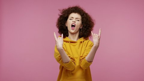 Disobedient excited girl afro hairstyle in hoodie feeling crazy, showing rock and roll hand gesture, punk sign, shouting and roaring wild on hard rock festival. studio shot isolated on pink background