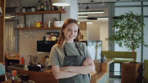 Portrait of happy woman standing in her cafe coffee shop. Cheerful young waitress waiting for clients at coffee shop. Successful small business owner in casual wearing grey apron standing at entrance
