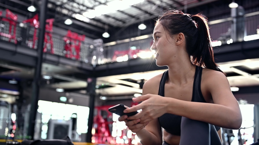 Sporty woman using mobile phone after workout. Asian woman exercise and lifestyle at fitness gym. Wellness and healthy for bodybuilding. | Shutterstock HD Video #1056839477