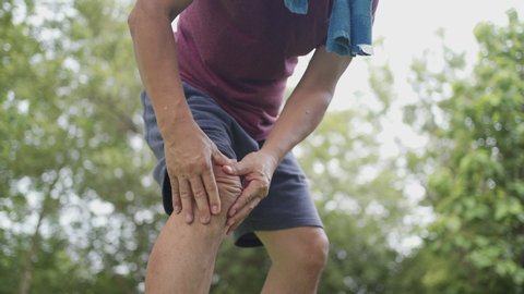 Asian tan skin male having painful knee injury during jogging exercise Inside the park with trees on the background, body condition knee pain, joint ligament problem, out door exercise knee ache