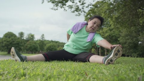 Asian active overweight mature woman doing split legs stretching hand to toe sit on grass lawn after exercise at the outdoor park, health insurance, human body flexibility, Legs muscle full extension