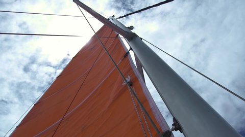 View of mainsail, mast and ropes from low angle with sun shining above, white cloud and blue skies in the background. Summer sailing concept.