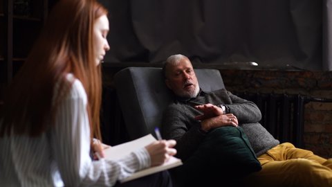 Depressed mature man lying on couch in counseling office and discussing troubles with compassionate female psychologist. Psychoanalyst listening older patient and writing notes in psychologist office