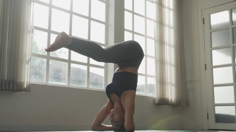 Asian young fit sport woman training yoga doing headstand pose on floor in room in evening. Girl concentrate on body strength and lift body up, beautiful posture. Meditation yoga workout for health.