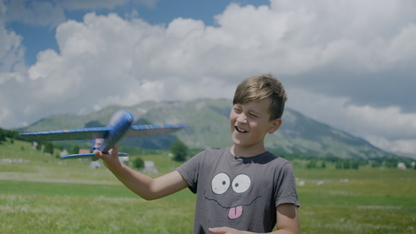 Happy boy kid plays with a toy blue plane on meadow with flowers against a background of mountains in Montenegro. Happy childhood concept. 
