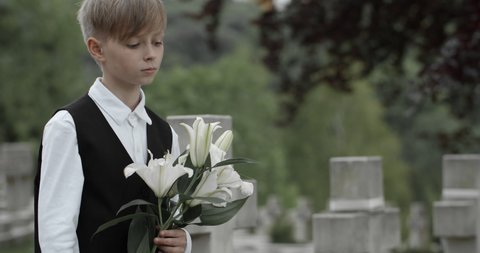Close up view of boy teenager putting white lily flowers on gravestone of his father. Young kid honoring his dad soldier at cemetery. Concept of memorial day.