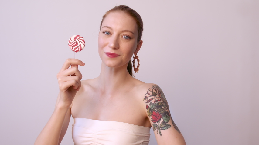Happy tattooed young woman holding swirl lollipop.Beauty female model with red hair style and beautiful trendy makeup holding pink sweet colorful lollipop candy