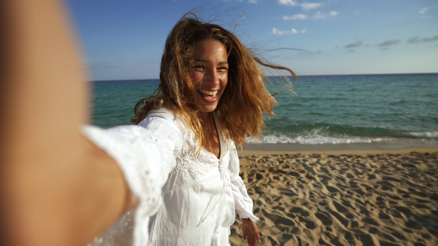 A happy carefree young female tourist with a straw hat is making selfie or technology video call with a phone to a friends or parents on a desert offshore seaside in a sunny day during vacation trip.