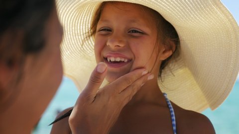 Close up of young mother is applying protective sunscreen or sunblock lotion on her little happy smiling daughter's face to take care of skin on a seaside beach during family holidays vacation.