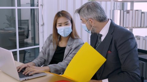 Normal view, mediup shot, 45 angle front view of professional caucasian boss explaining work to young asian female secretary in suit, both wearing facial maskin the office during DOVID-19 pandemic.