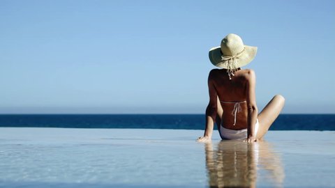 Sexy woman in summer hat and bikini sitting in infinity pool and taking sunbath