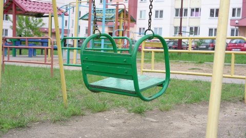 vintage metal wooden green swing sways on rusty chains against playground attractions and building slow motion