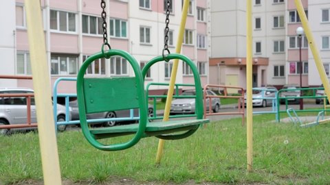 old green swing with brown rusty chains on playground against parking lot and local building slow motion close view