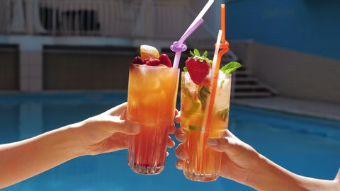 Close up view of young female hands holding exotic non-alcoholik ice fruit cocktails, making cheer, clink glasses, joy drinking and celebrate friendly meeting near pool in luxury spa complex.