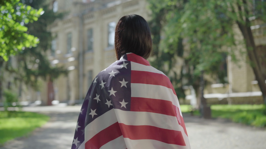 Back view of brunette woman in American flag making liberty hand gesture. Young slim lady showing freedom sign at university yard. Concept of independence or protest.