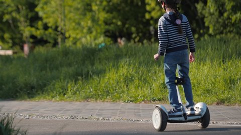 KAZAN, TATARSTAN/RUSSIA - MAY 28 2020: Little girl in helmet rides modern hoverboard during mother helps son to use scooter on road in green park on May 28 in Kazan