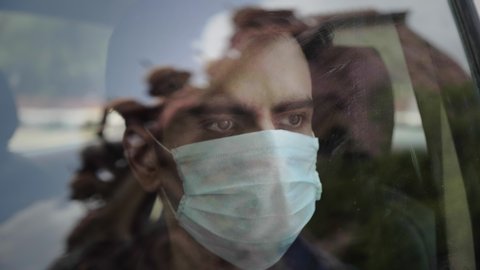 A man male sitting in the car wearing a face protective mask looking outside through the glass window amid Corona virus COVID 19 epidemic or pandemic