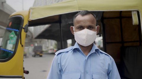 An man or male auto rickshaw or tuk tuk driver looking at the camera standing outdoors along the street wearing face protective mask amid Corona virus Covid 19 epidemic or pandemic