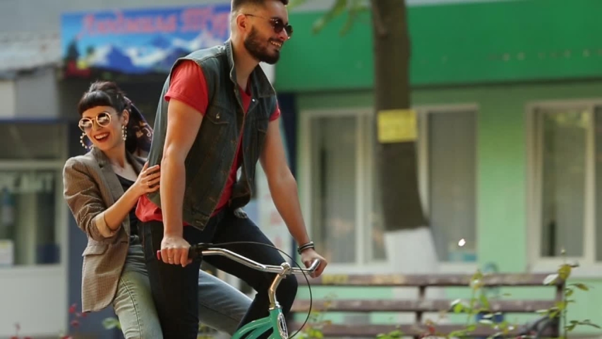 Young couple riding on bicycle together in the park. Romantic boyfriend cycling with happy girlfriend on cargo rack on city street. Woman hugs man having fun riding on bike in tandem. Slow motion. | Shutterstock HD Video #1056899006