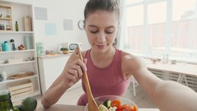 Medium shot of young Caucasian sporty woman sitting in the kitchen, holding camera in her hand and telling about healthy nutrition