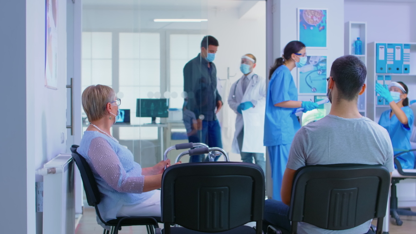 Private hospital reception area during global pandemic. Doctor with visor against coronavirus inviting man in his office for examination. Disabled senior woman with walking frame | Shutterstock HD Video #1056901550