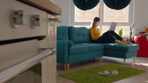 A young woman sits on sofa in co-working space. The girl in medical mask and uses laptop. Sunny room with large windows. Video de stock