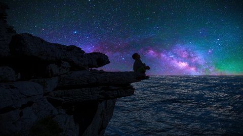 The silhouette of a boy sitting on a cliff edge watching the milky way and shooting stars.