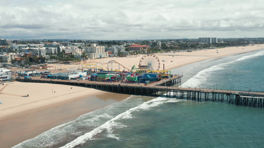 Santa Monica Pier on cloudy day, Los Angeles. The footage of the Santa Monica pier and the amusement park from the side of the ocean. Beautiful panorama of the city skyline is seen behind sandy beach Royalty-Free Stock Footage #1056903743