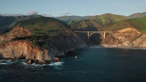 Outdoor travel. Panoramic aerial over the rocky ocean coast at sunset. Beautiful ocean water meets the rocky shore. Scenic arch bridge at the pure nature of Big Sur. 4K mountains on background, USA