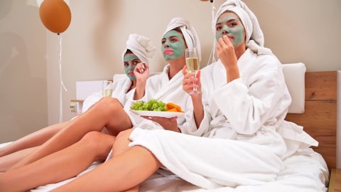 Side motion of group of girls with cosmetic masks, in towels and bathrobes enjoying home party. Three female friends holding glasses with champagne, eating fruits and watching movie in bedroom.