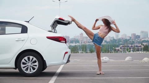 Beautiful Slim Woman in short denim shorts sexually goes to her car and opens the trunk to put on a hat. Athletic girl with a perfect figure and a steep stretch.