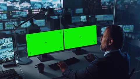 Male Officer Works on a Computer with Two Green Screen Mock Ups in Surveillance Center in a Harbour Monitoring Center with Multiple Cameras on a Big Digital Screen. Employees Sit in Front of Displays.