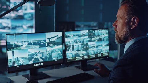 Male Officer Works on a Computer with Surveillance CCTV Video Footage in a Harbour Monitoring Center with Multiple Cameras on a Big Digital Screen. Employee Uses Radio to Give an Order or Report.