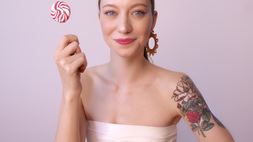 Happy young woman holding colorful sugar candy, over background.Beauty tattooed female model girl with glowy makeup and red lips with big swirl lollipop in her hand