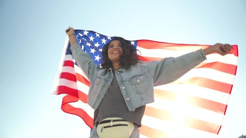 Happy young African woman holding usa United States flag dancing outdoor under sunny sky. Smiling free proud independent patriotic Afro American girl feeling freedom, independence concept. Slow motion