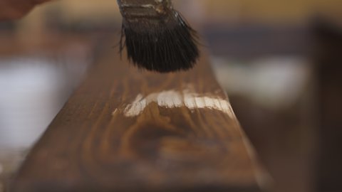 Slow-motion close up of man carpenter painting a piece of wood with a brush. Painting with brown stain primer a floorboard. DIY.