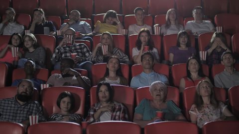 Scared multinational audience watching horror in movie theater. Viewers covering their eyes with fear. Multi-ethnic theater-goers enjoying scary movie in cinema