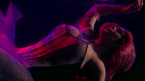 a woman with long hair and a corset hovers horizontally under the surface of the dark water and touches it with her hand. purple contour light
