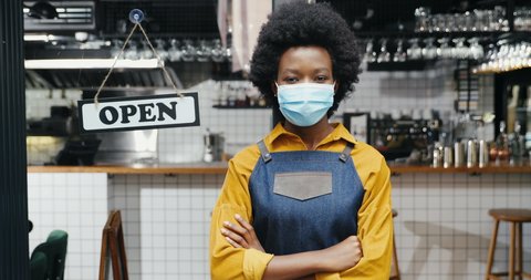 Portrait of beautiful African American woman waitress in apron and medical mask looking straight with table open at cafe entrance outdoor. Pretty barista at bar door with board open Reopen Zooming in