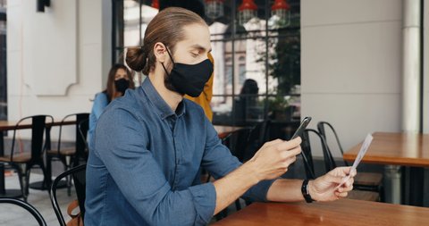 Caucasian man client in mask siting at table in cafe outdoor and scanning QR code with smartphone for reading menu. Technologies during covid-19 pandemic. Male customer in bar making order on phone.