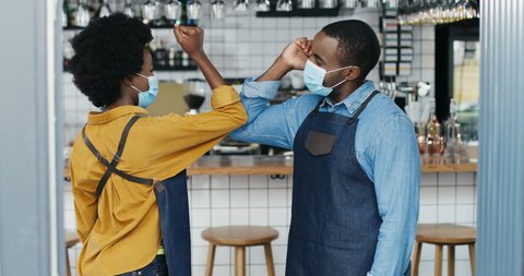 African American young cheerful waiter and waitress in masks, aprons and gloves laughing and touching with elbows like greeting in cafe. Quarantine reopen concept. Male and female baristas meeting.