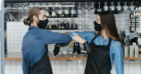 Caucasian young cheerful waiter and waitress in masks, aprons and gloves laughing and touching with elbows like greeting in cafe. Quarantine reopen concept. Male and female baristas meeting in bar.